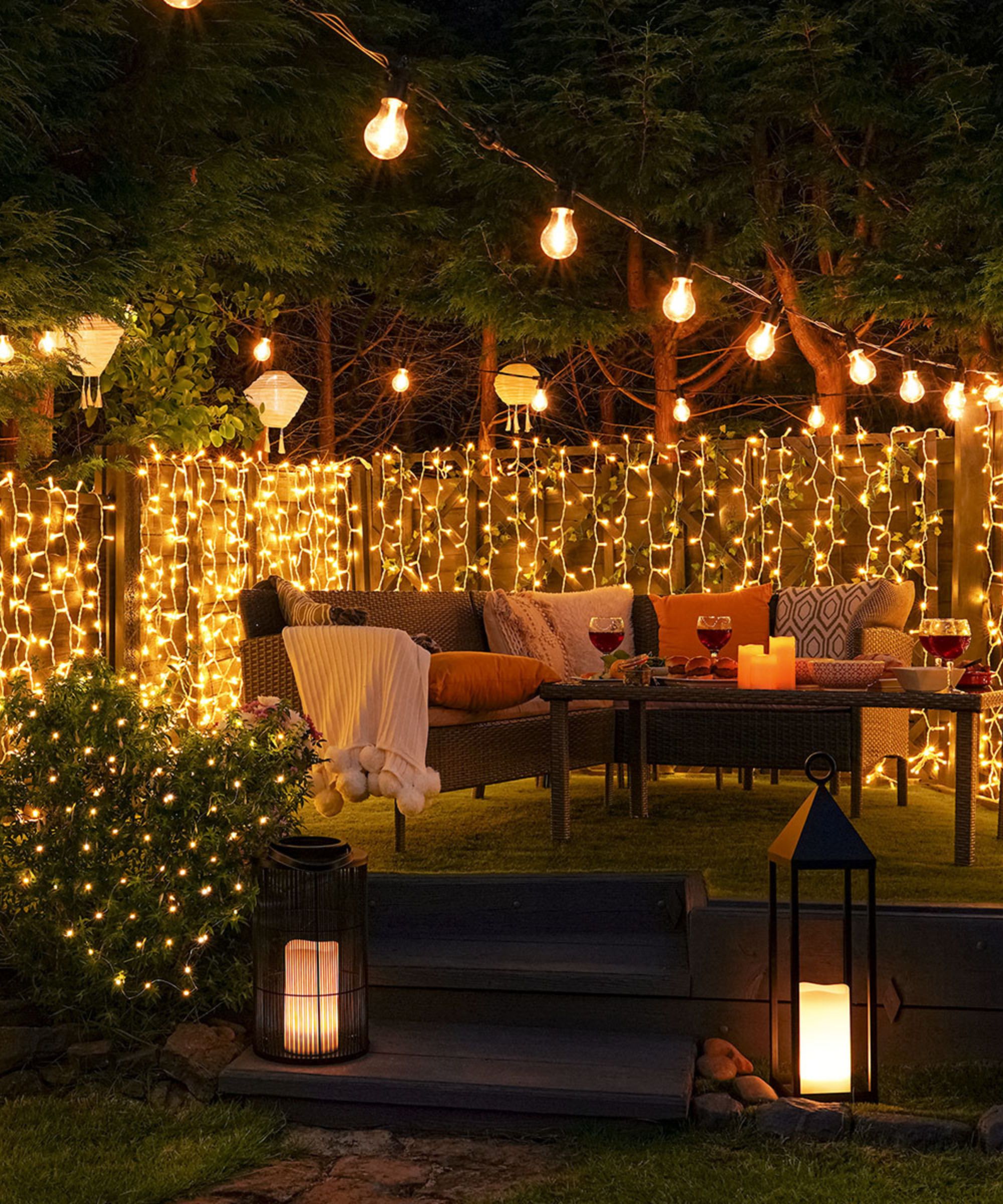 outdoor sofa with cushions and blankets lit up by festoon lights, string lights and hurricane lanterns