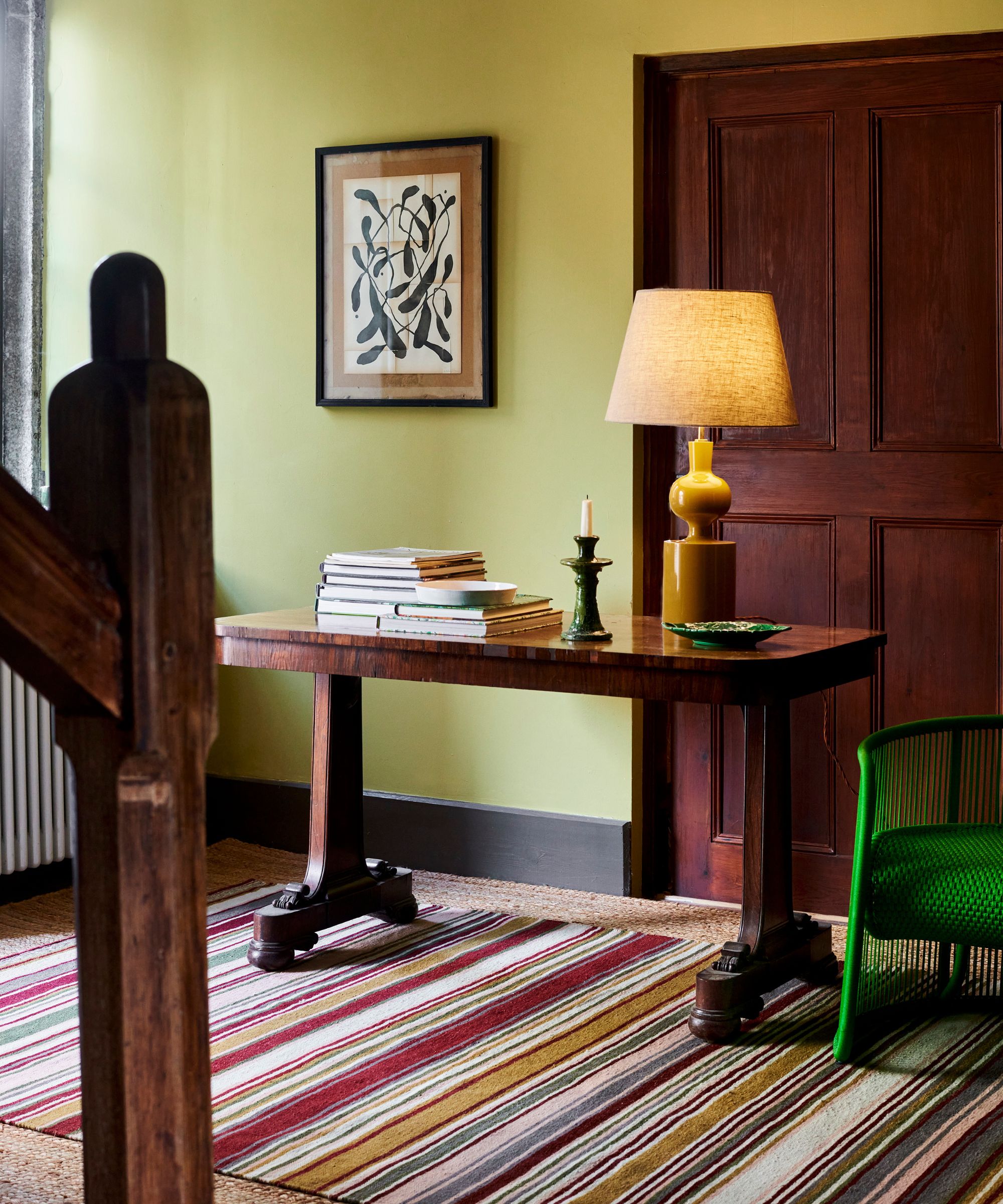 Hallway with striped colourful rug, green chair, console with books and yellow lap and staircase