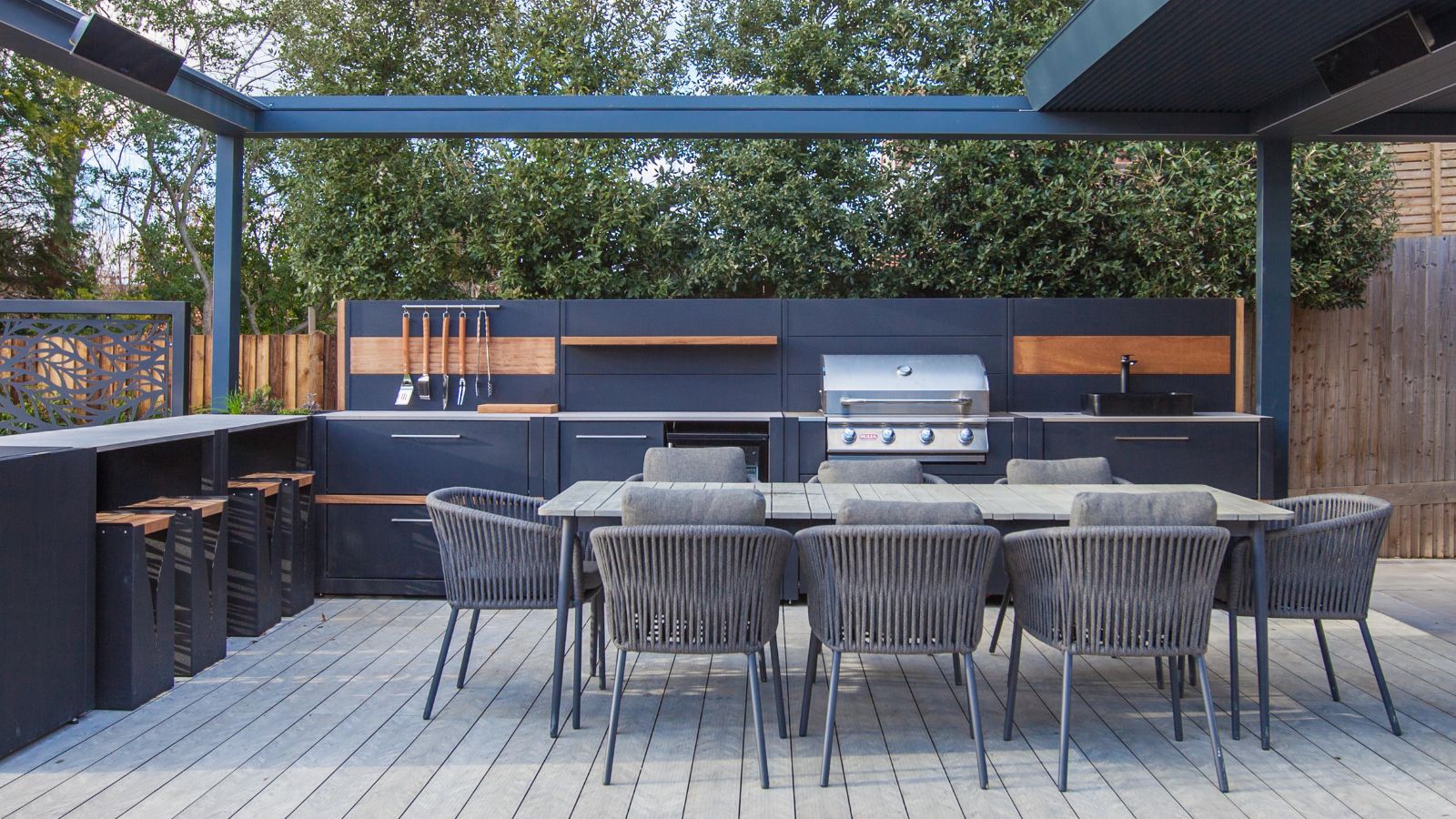 A large outdoork itchen area with grey metal cabinets and backsplash, and a built in grill, behind a grey roped dining set
