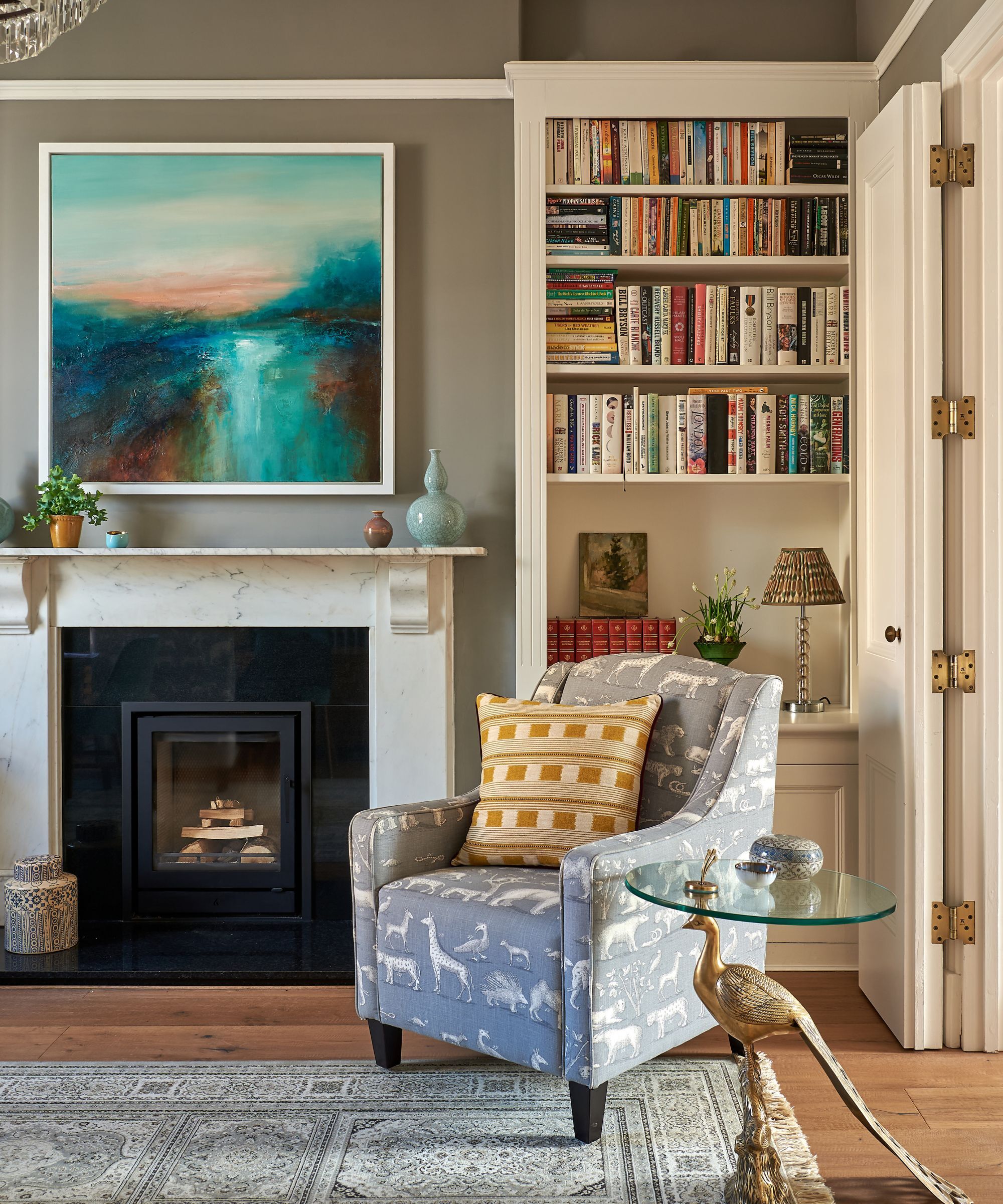 A sitting room with a wood burning stove, wooden floors, gray rug, gray armchair with a yellow cushion, a glass side table and behind an inbuilt bookcase. The walls are painted in Portland Stone by Little Greene